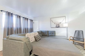 A room with a large bed, a sofa, a chair, and a wall-mounted lamp.