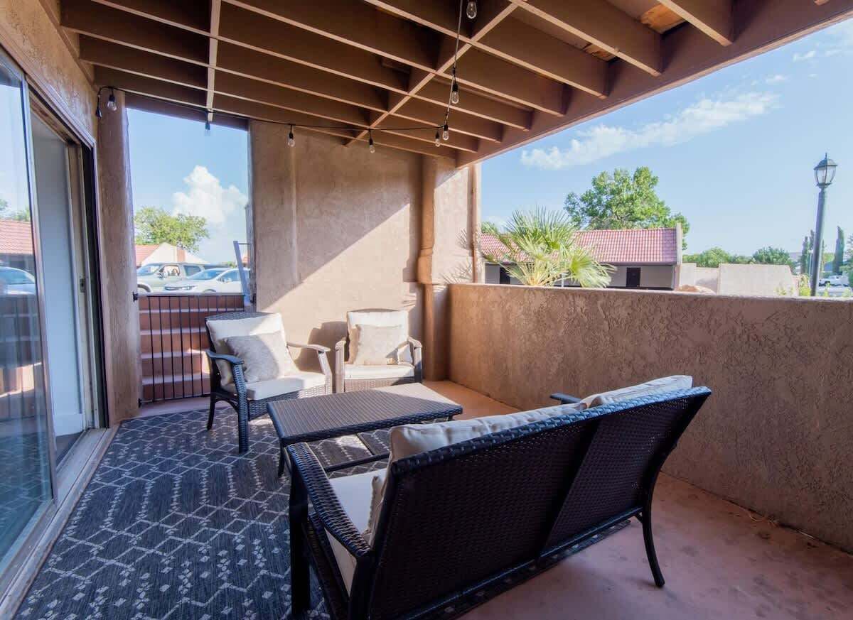 An outdoor balcony with wicker furniture, a glass door leading to a parking area, and a view of the surrounding neighborhood.