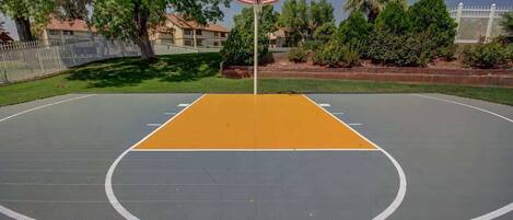 An outdoor basketball court with a hoop, surrounded by trees and a fence.