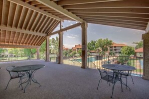 An outdoor patio area with two tables and chairs, a covered roof, and a view of a swimming pool and buildings in the background.