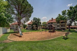 Un parque infantil con columpios y un tobogán, rodeado de césped y árboles, con edificios al fondo.