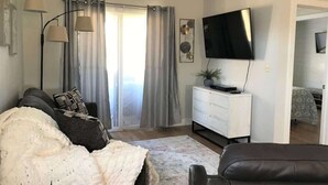 A living room with a sofa, a television, a white dresser, and a large window with curtains. - Sports Village Resort #H9 | All Amenities Included (Saint George)