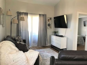 Ein Wohnzimmer mit einer Couch, einem Fernseher, einer weißen Kommode und einem großen Fenster mit Vorhängen.