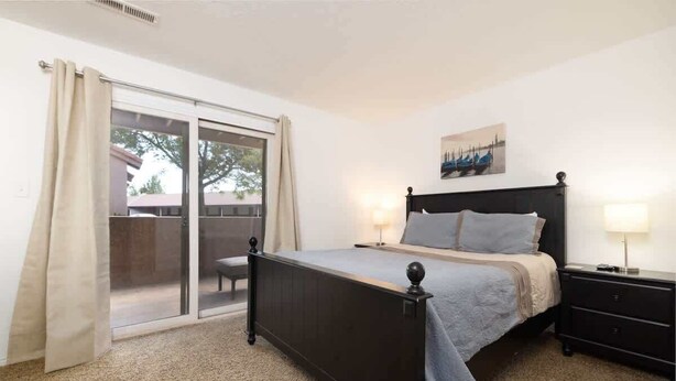 A bedroom with a bed, a nightstand, a lamp, a window with curtains, and a view of the outside.