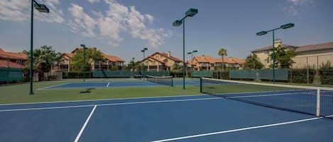 Een tennisbaan met meerdere velden, omringd door gebouwen met rode daken en groene omheining.