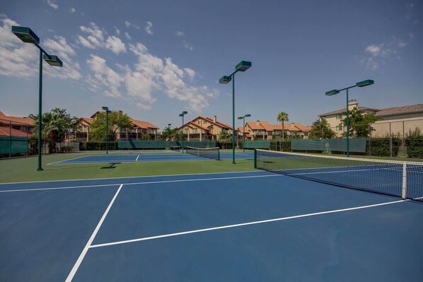 A tennis court with multiple courts, surrounded by buildings with red roofs and green fencing.