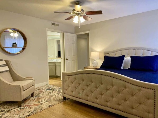 A bedroom with a tufted headboard bed, a round mirror, a chair, and a ceiling fan.