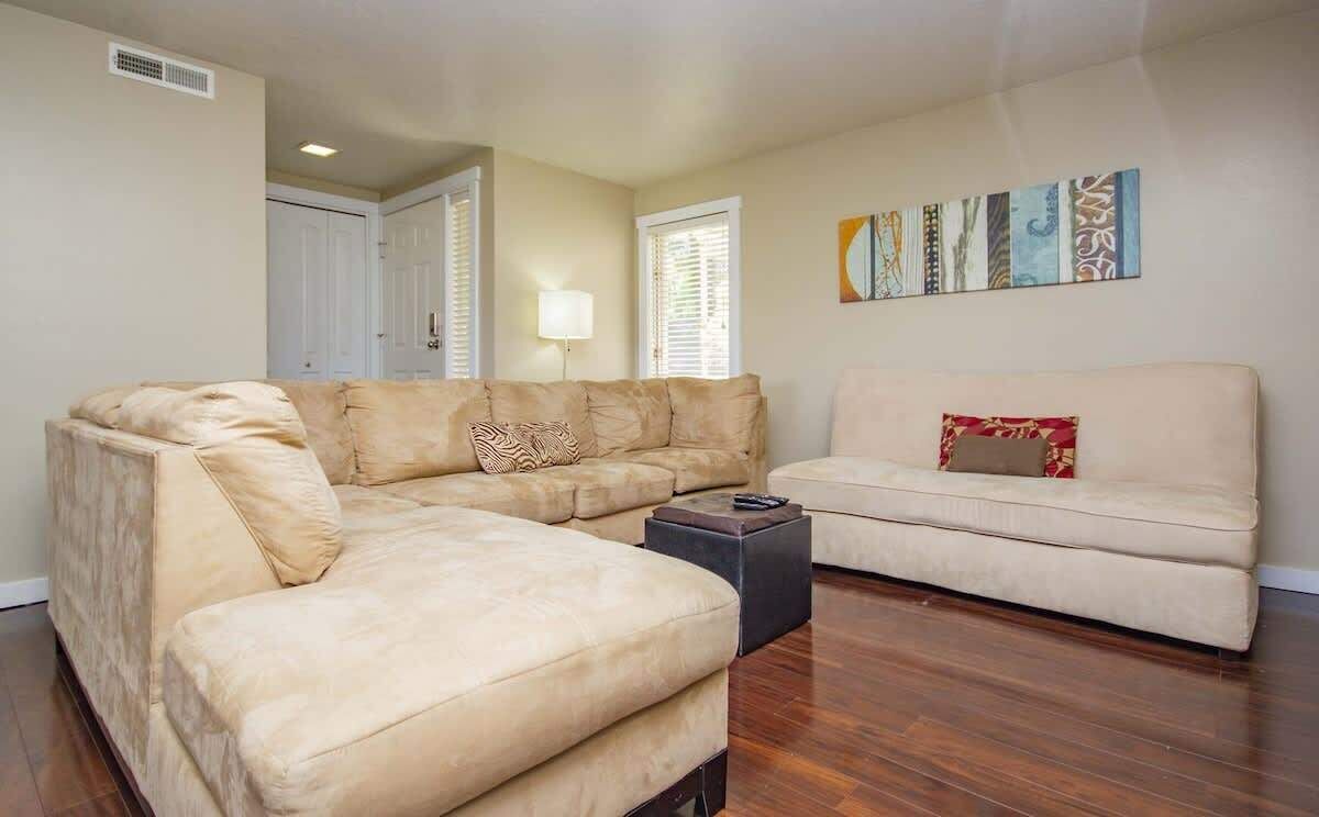 A living room with a beige sectional sofa, a smaller sofa, and a coffee table. There is a door and a window with blinds in the background.