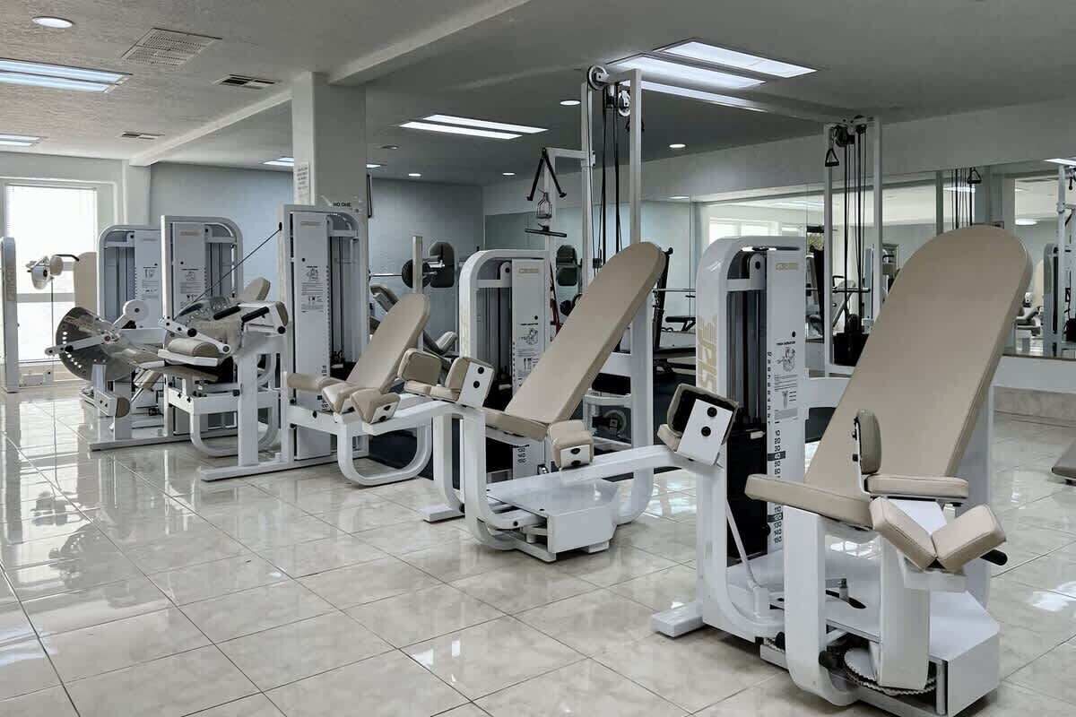 A gym with various exercise machines, including weight benches and cardio equipment, arranged in a spacious room with a tiled floor and large windows.