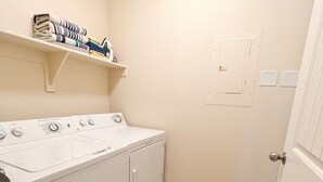 A laundry room with a front-loading washer and dryer, a shelf with folded towels, and a door with a light switch.