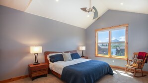 Un dormitorio con cama, dos mesitas de noche con lámparas, un sillón mecedora y un ventanal con vistas a edificios y montañas.