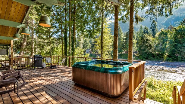 En træterrasse med jacuzzi, udendørs møbler og udsigt til en flod og skov.