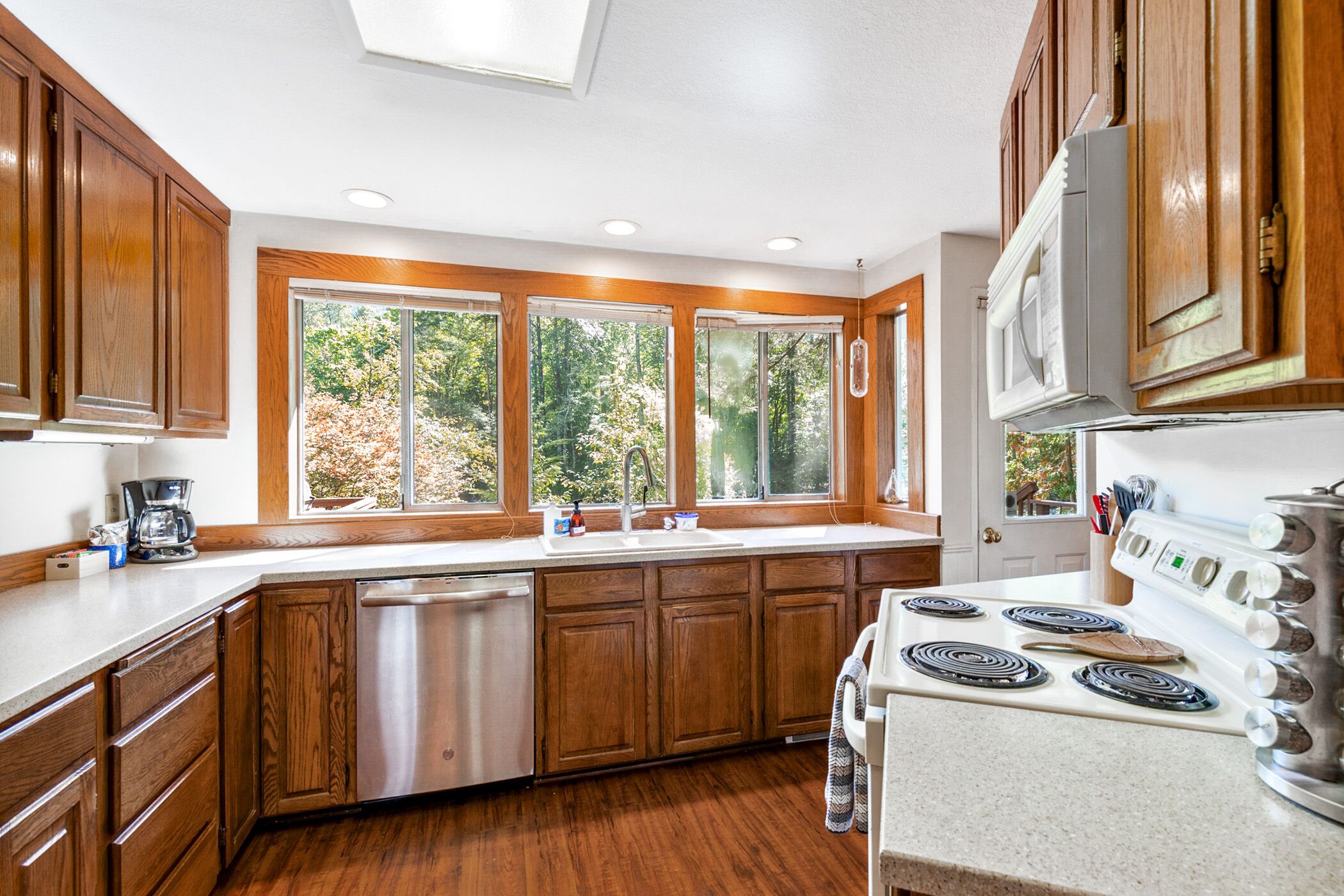 Una cocina con armarios de madera, lavavajillas de acero inoxidable, pileta doble, estufa y ventanas amplias con vista a árboles.