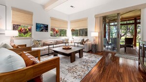 A spacious living room with a large sofa, a coffee table, and a dining area with wicker furniture. There are large windows with blinds and a view of greenery outside. - 🏖️Kohala Coast Townhome with Beach Club Access | Mauna Lani (Kamuela)