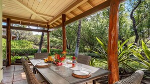 An outdoor covered patio with a wooden table set for dining, surrounded by wicker chairs, and a lush garden view. - 🏖️Kohala Coast Townhome with Beach Club Access | Mauna Lani (Kamuela)