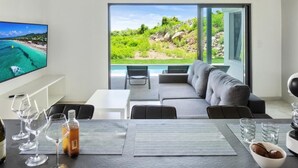 Una sala de estar moderna con un televisor grande, una mesa de comedor con vasos y una botella, y vistas a una piscina y vegetación.