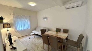 Ein modernes, helles Zimmer mit Esstisch, Stühlen, einer Couch und einem kleinen Schreibtisch mit Lampe und Pflanze.
