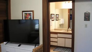 Ein Zimmer mit einem Flachbildfernseher auf einer Kommode, ein Badezimmer mit Waschbecken und Spiegel sowie eine Tür zu einem weiteren Raum.