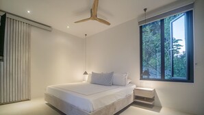 Un dormitorio moderno con una cama grande, una mesita de noche, un cabecero de madera, una ventana con vistas a árboles y un ventilador de techo.