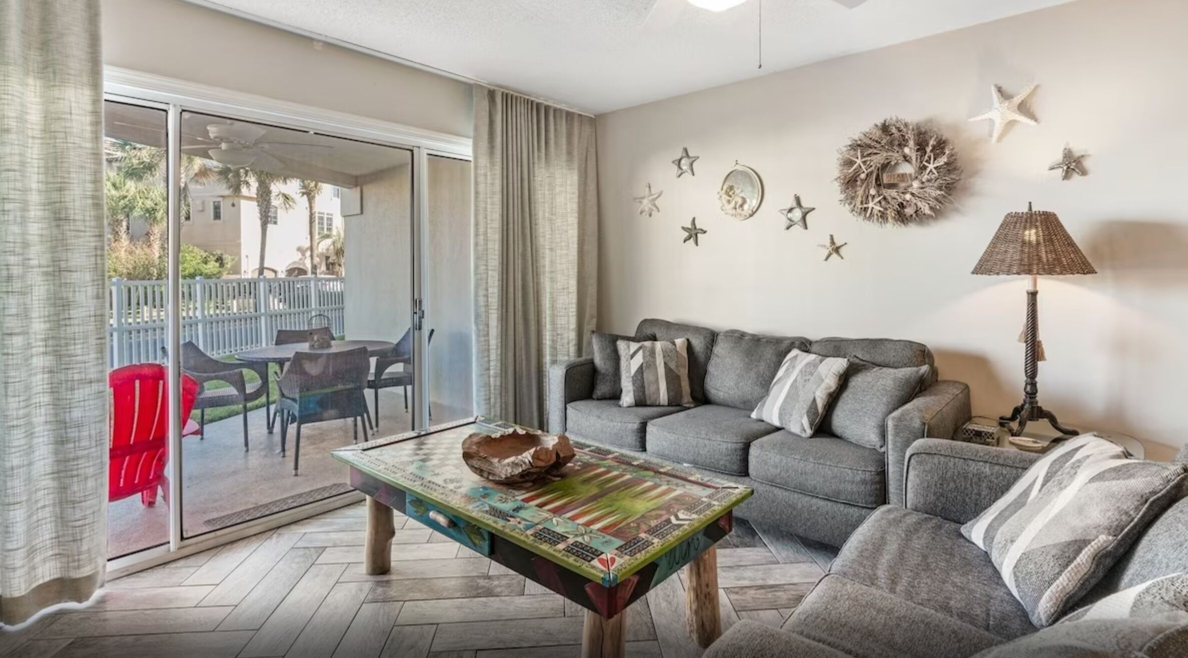 Ein Wohnzimmer mit grauem Sofa, einem Couchtisch mit dekorativer Schale und einer Lampe. Sterne sind an der Wand angebracht und es gibt einen Blick ins Freie durch eine Schiebetür.