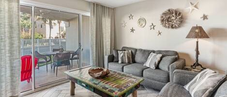 A living room with a grey sofa, a coffee table with a decorative bowl, and a lamp. There are star decorations on the wall and a view of the outside through a sliding door.