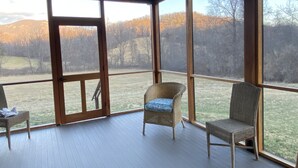 Une véranda couverte avec deux fauteuils en osier et une vue sur un champ verdoyant et des arbres.