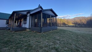 Un bâtiment à deux étages, doté d’un porche abrité et de grandes fenêtres, situé dans un champ verdoyant avec des montagnes en arrière-plan.
