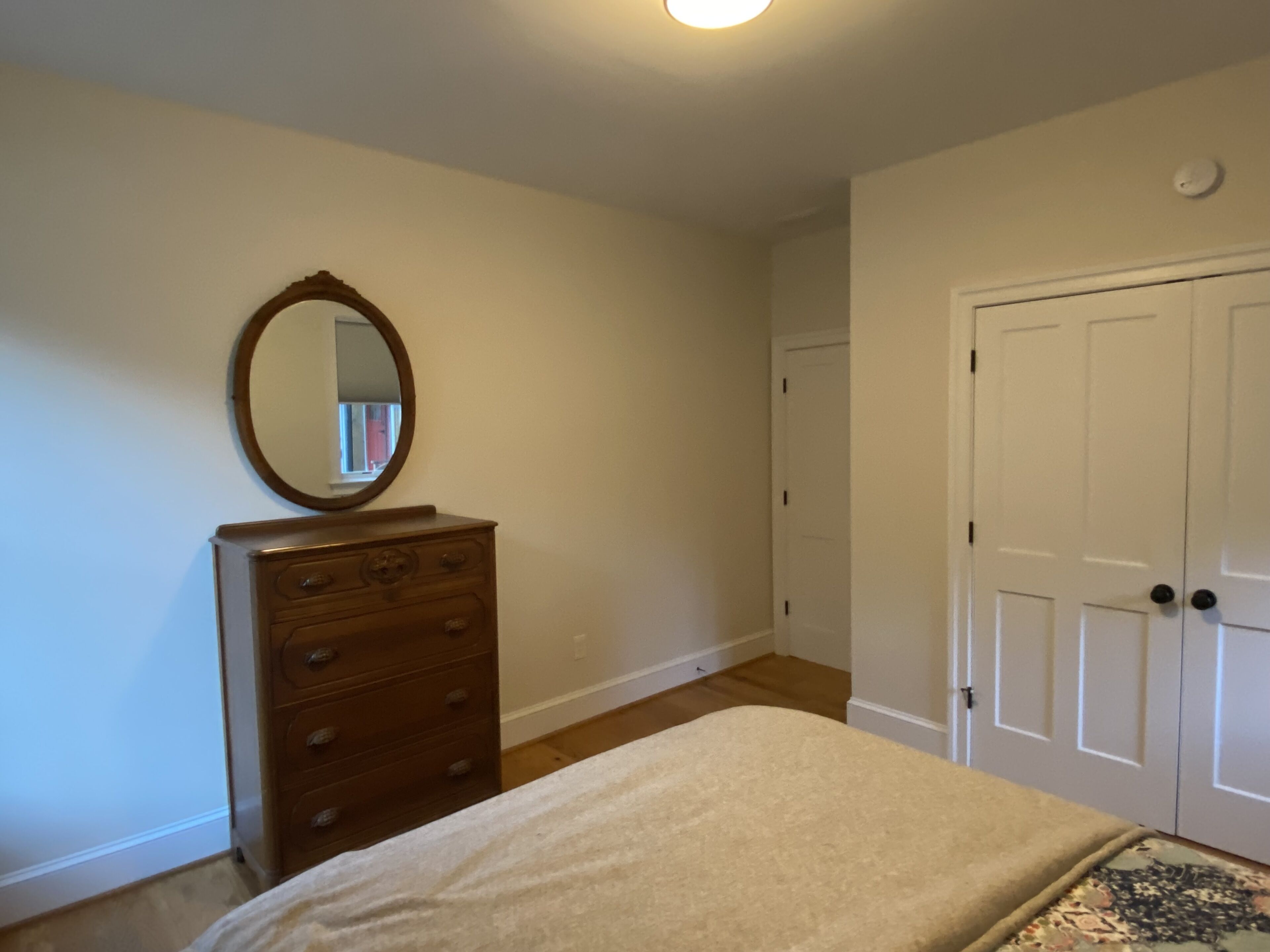 A bedroom with a wooden dresser, a bed with a beige blanket, a round mirror, and a white door.