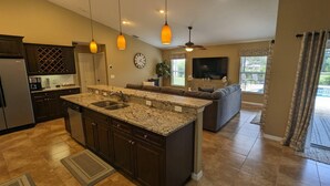 A modern kitchen with granite countertops, dark cabinetry, and a built-in island with a sink. There is a dining area with a sofa and a television.