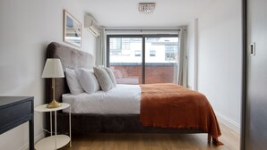 A modern bedroom with a large window, a bed with a brown headboard, a bedside table with a lamp, and a view of a brick building outside.