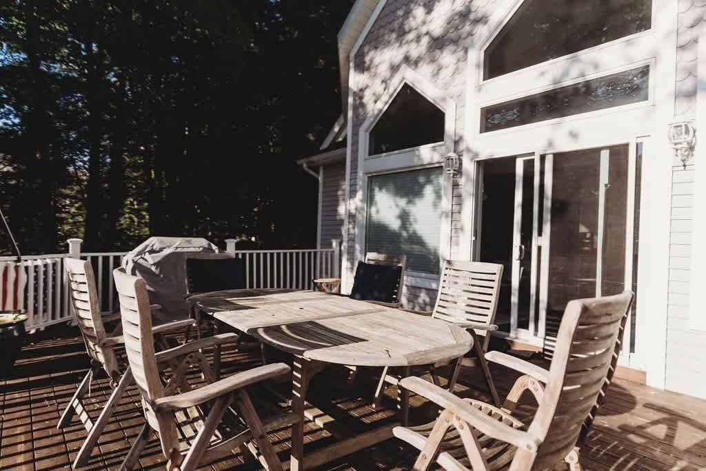 Une terrasse en bois avec une table ronde et des chaises, un barbecue et une maison aux grandes fenêtres et à l’extérieur blanc.
