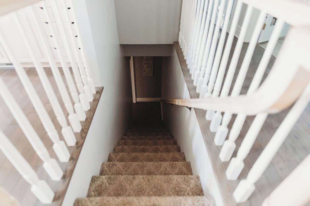 Uma escadaria com balaústres brancos e degraus carpetados, que dá acesso a um patamar.