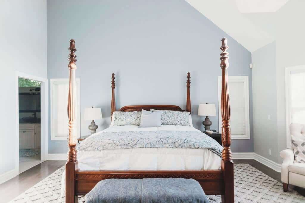 A four-poster bed with a wooden frame and a matching bench, set in a room with light blue walls and a large window.