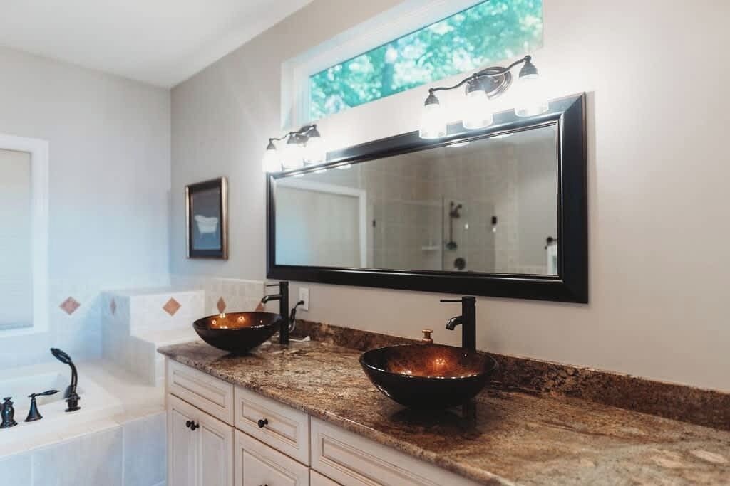 A bathroom with a large mirror, two vessel sinks, a bathtub, and a window with a view of trees.