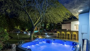 Un bain à remous à l’intérieur bleu, entouré d’un jardin aménagé avec des arbres et des arbustes. On y trouve également un espace patio avec des chaises jaunes et une table.