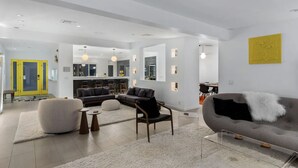 Un salon moderne avec un canapé gris, un fauteuil noir et un pouf rond. On y trouve également un coin cuisine avec un bar et des tabourets.