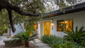 Une maison moderne avec une porte jaune, entourée d’un jardin où poussent différentes plantes et un grand arbre.