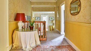 Un couloir avec un papier peint à motifs, un lustre, une table ronde avec une lampe et un miroir au mur.