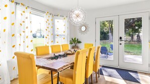Un coin repas avec une table en bois et des chaises jaunes, une plante en pot, et une vue sur l’extérieur grâce à une porte vitrée.