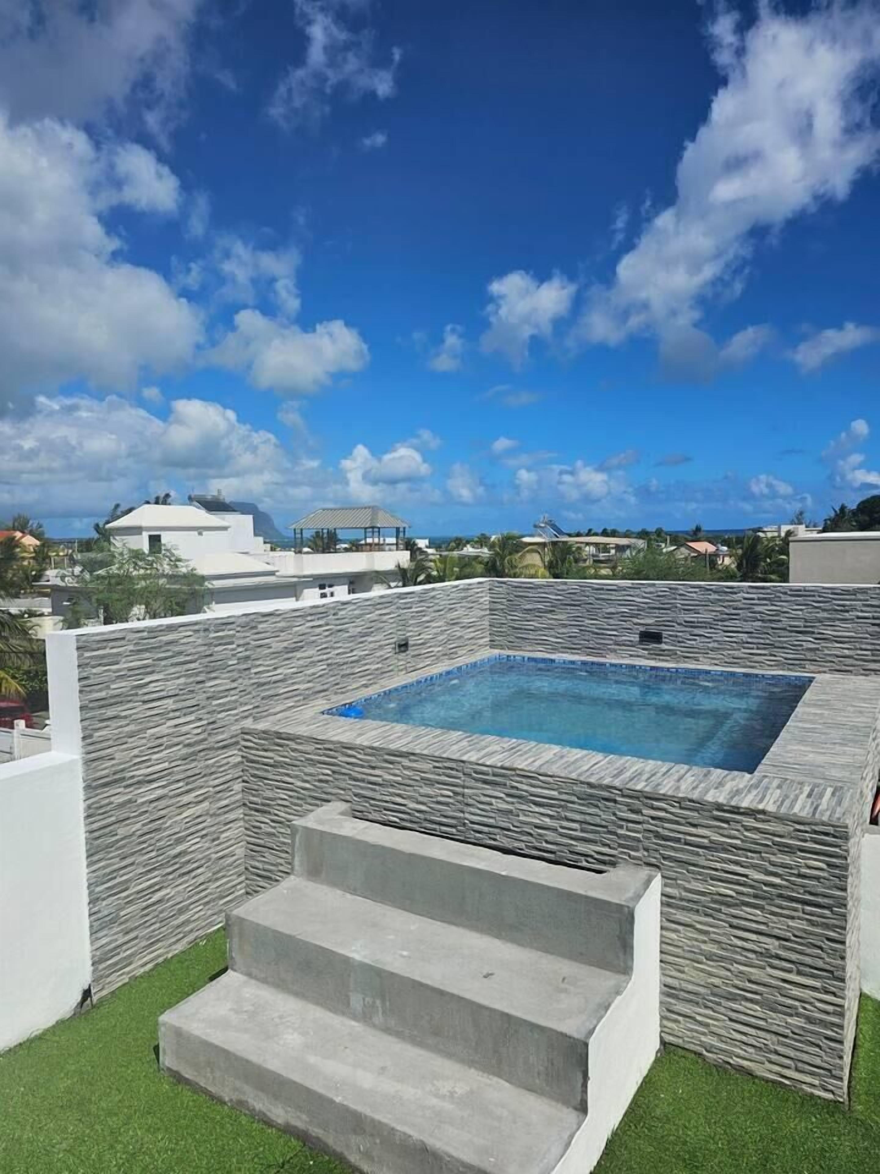En pool på taget med stenvægge, en lille trappe af beton op til poolen, og en klar blå himmel i baggrunden.