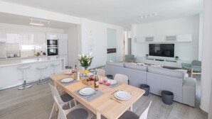 A modern kitchen with white cabinets, a dining area with a wooden table set for four, and a grey sofa in the living area. - Protaras Olivine Villa OL9 - Beautiful 4BDR Protaras Villa with Private Pool (Protaras)