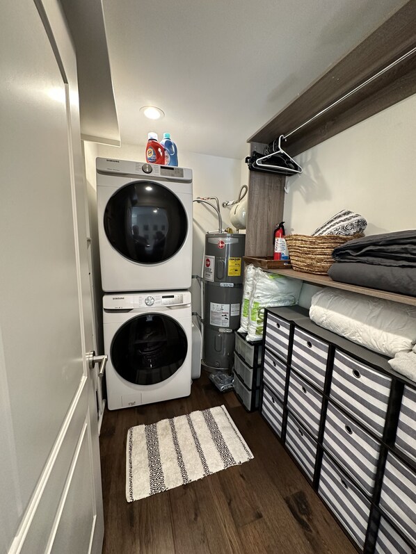 A compact laundry area with two Samsung washing machines, a water heater, and storage shelves.