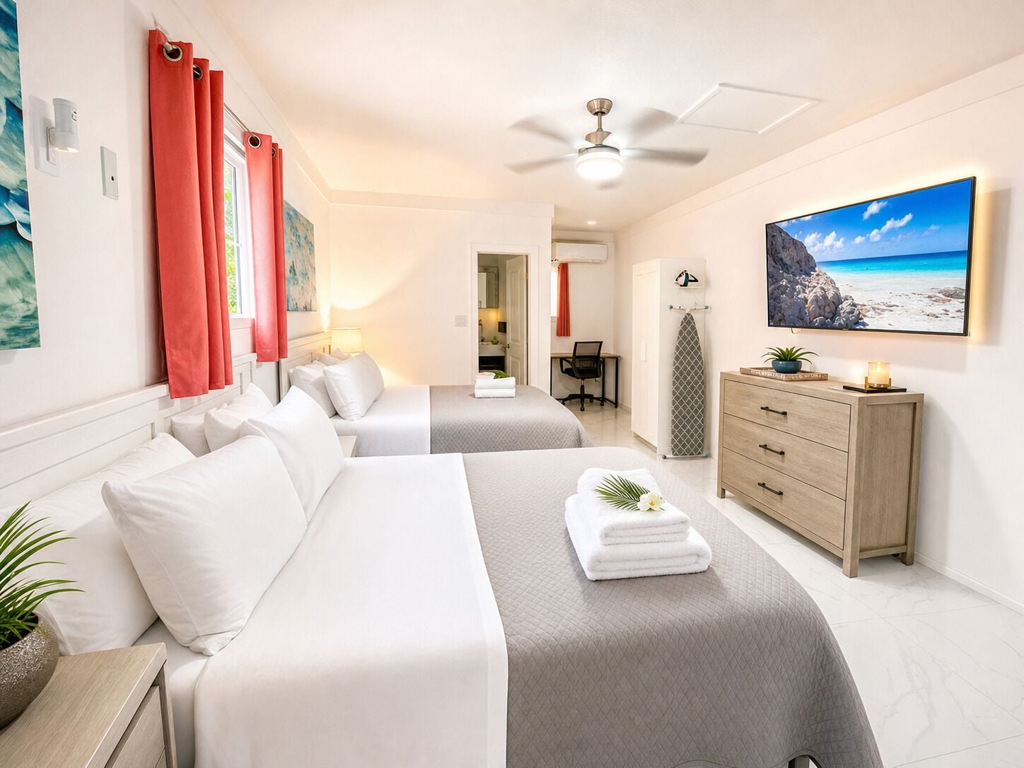 Hotelkamer met twee bedden, een bureau, een dressoir, een plafondventilator en een groot raam met uitzicht op het strand.