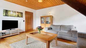 Een woonkamer met een flatscreen televisie, een houten salontafel, een grijze bank, een boekenkast en een houten plafond.