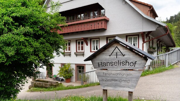 Een houten bord met de tekst 'Hanselishof Ferienwohnungen Zimmer 2/3' voor een gebouw met een balkon en een trap.