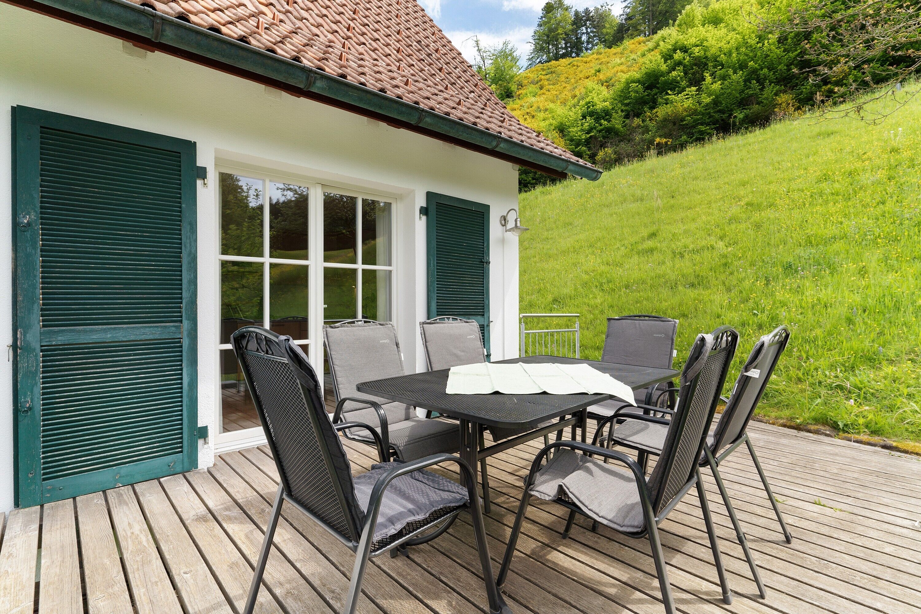 Eine Terrasse mit Tisch und Stühlen, ein weißes Gebäude mit grünen Fensterläden und ein grasbewachsener Hügel im Hintergrund.