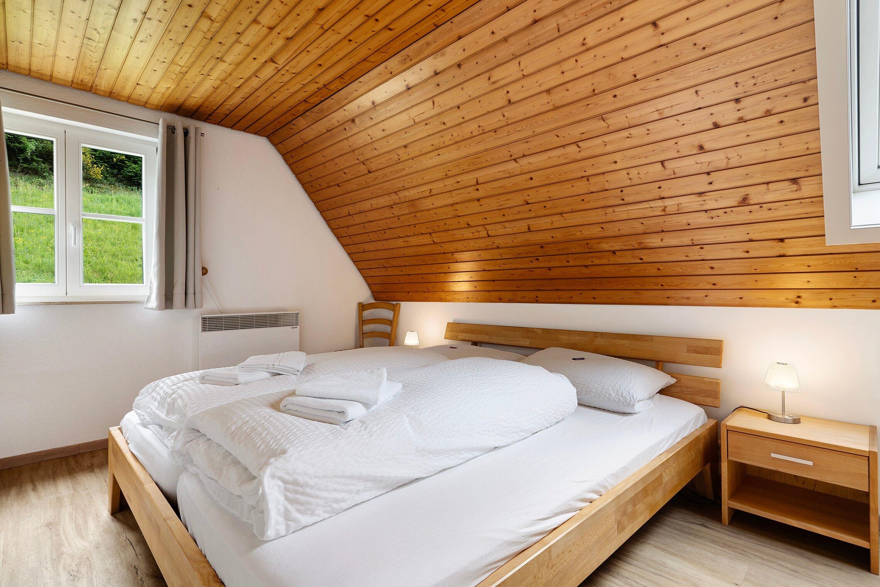 A bedroom with a wooden ceiling, a bed with white bedding, a nightstand with a lamp, and a window with curtains.