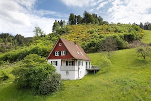 Uma casa com telhado vermelho e paredes brancas, cercada por vegetação exuberante e árvores.