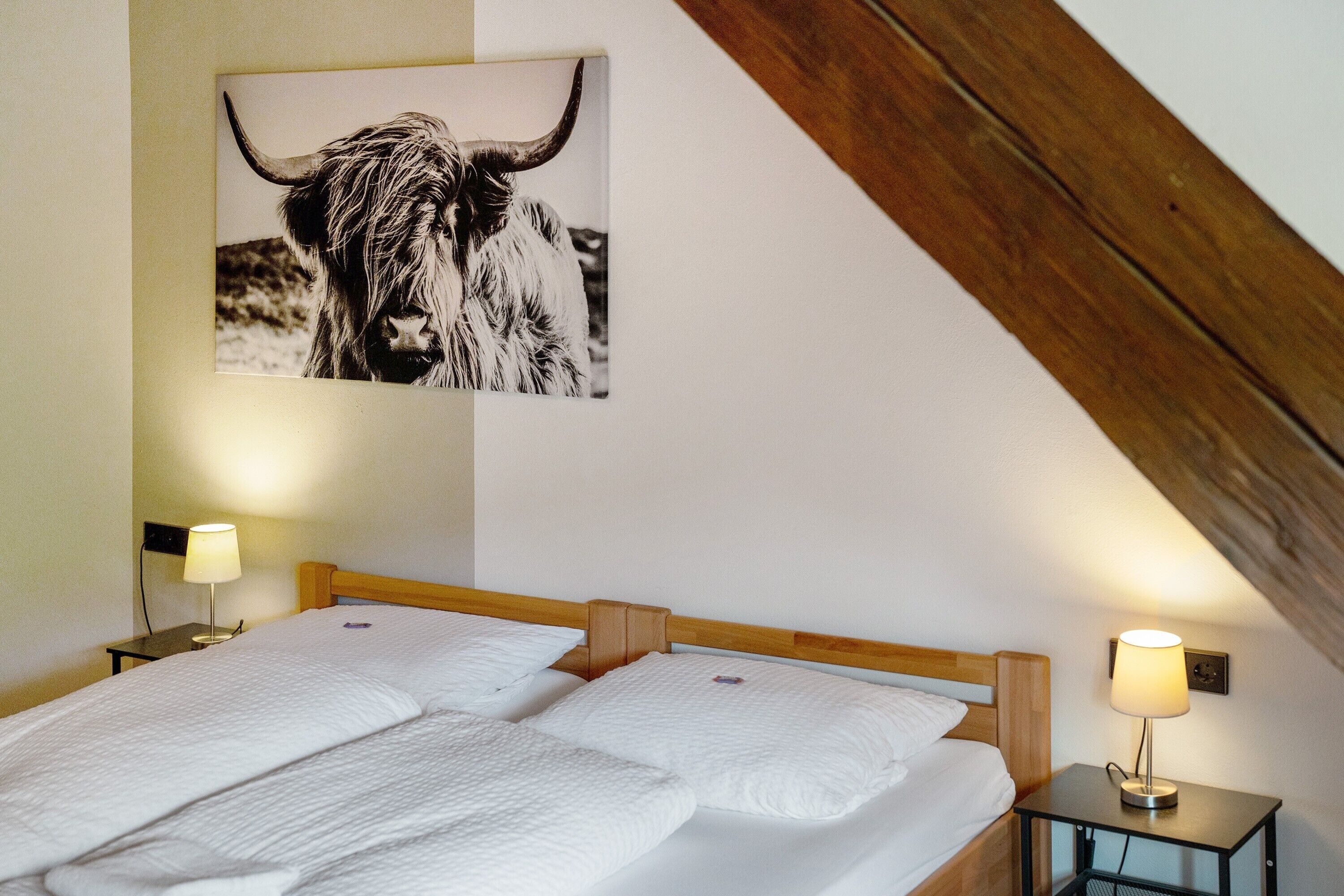 Une chambre avec deux lits, une tête de lit en bois, des tables de chevet avec des lampes et un tableau représentant un yak accroché au mur.