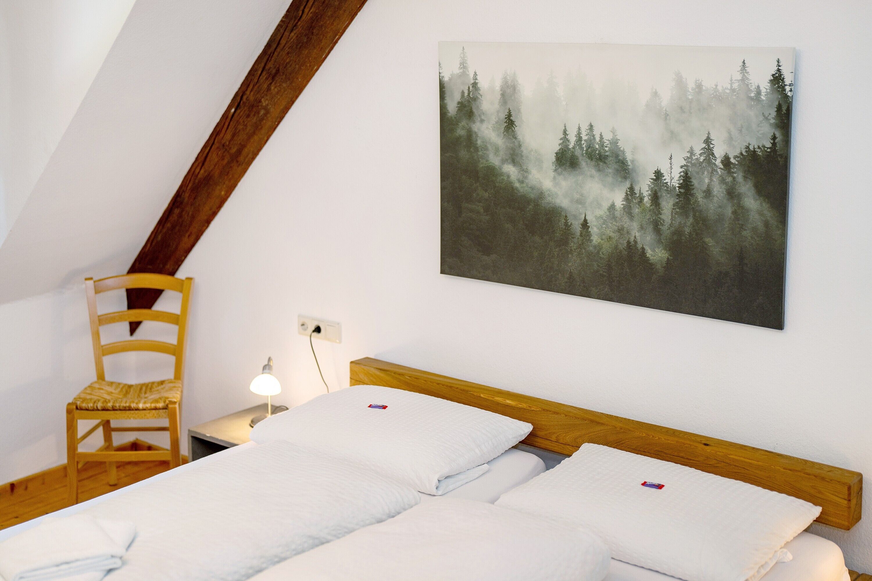 Un dormitorio con cama, una silla, una mesita de noche, una lámpara y un cuadro con motivos forestales en la pared.